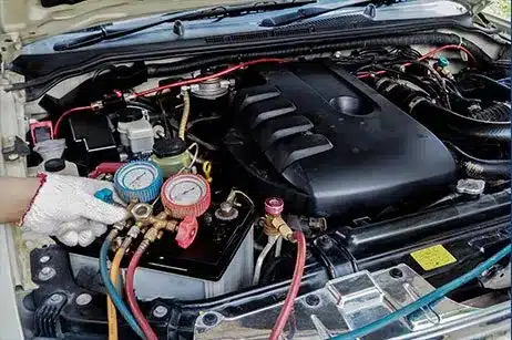 image of mechanic wearing white gloves hand, holding an auto air conditioner tester in front of engine on car with hood open.