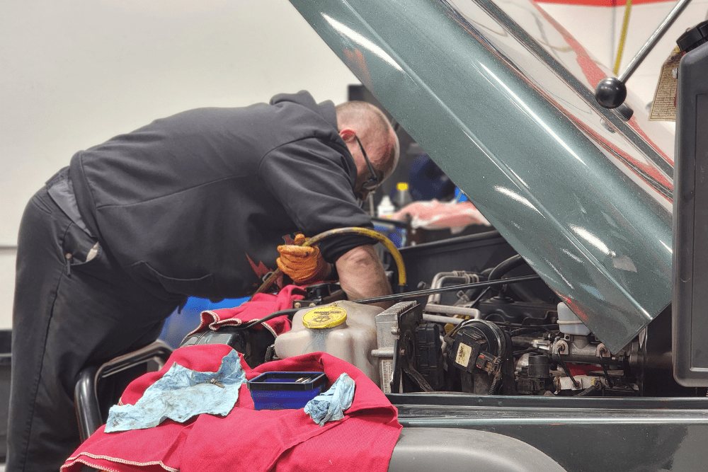 car repair, auto repair in Chandler, AZ at Mac's Complete Auto Repair. Auto technician working under the hood of a vehicle in a repair shop, using diagnostic tools and wearing gloves with red and blue shop towels nearby.